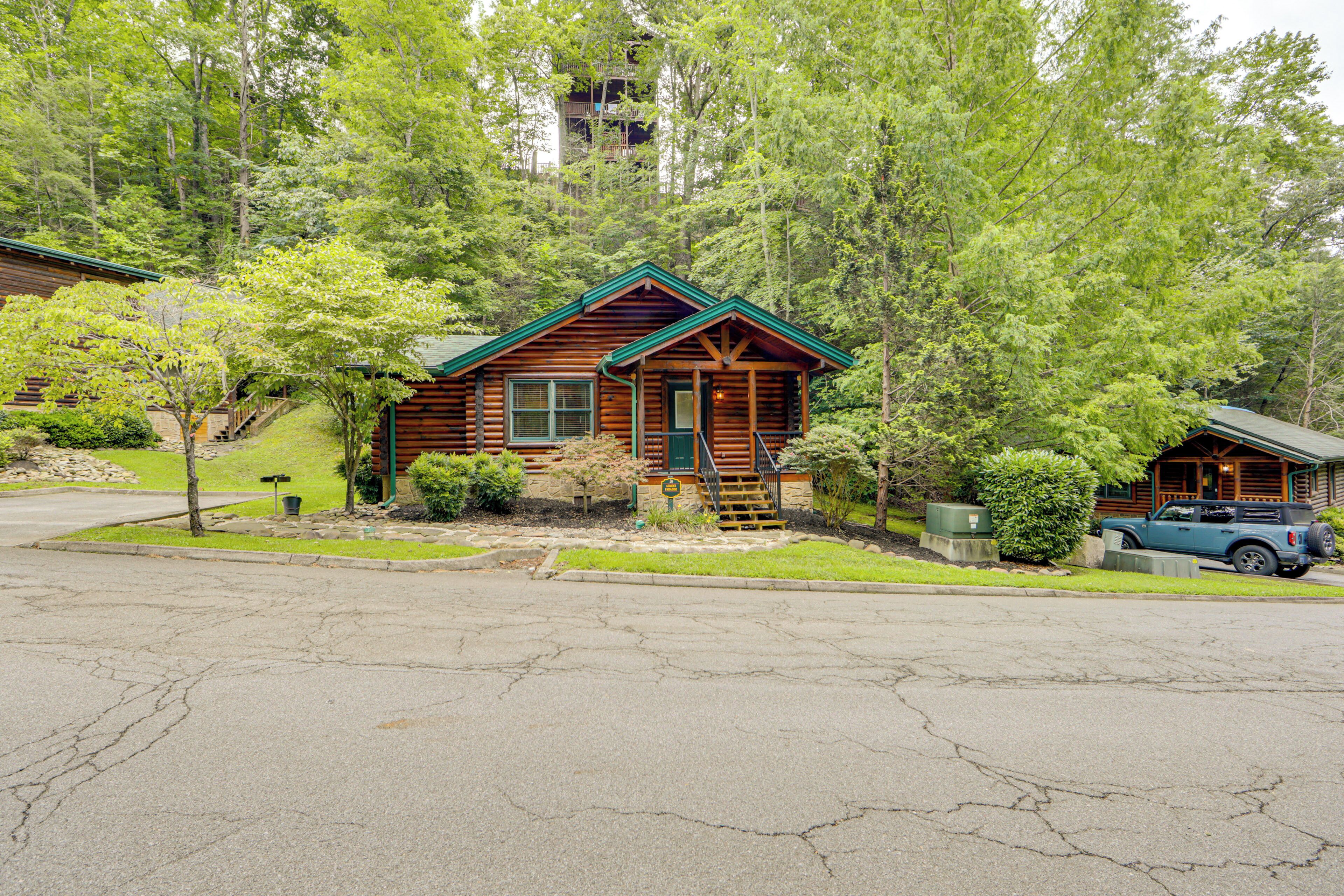 A log cabin with a green roof, surrounded by trees and a driveway with a parked car.
