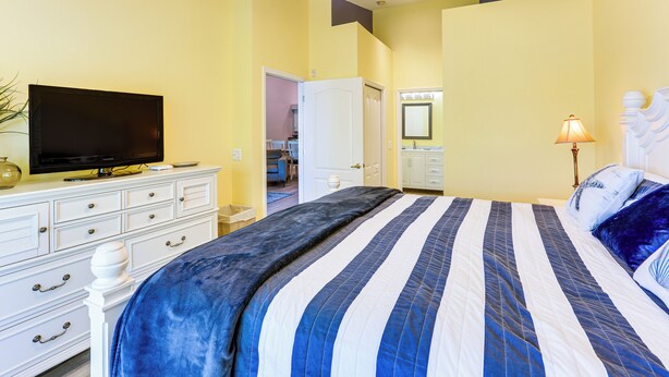A bedroom with a bed, a television on a dresser, a bathroom with a mirror, and a door leading to another room.