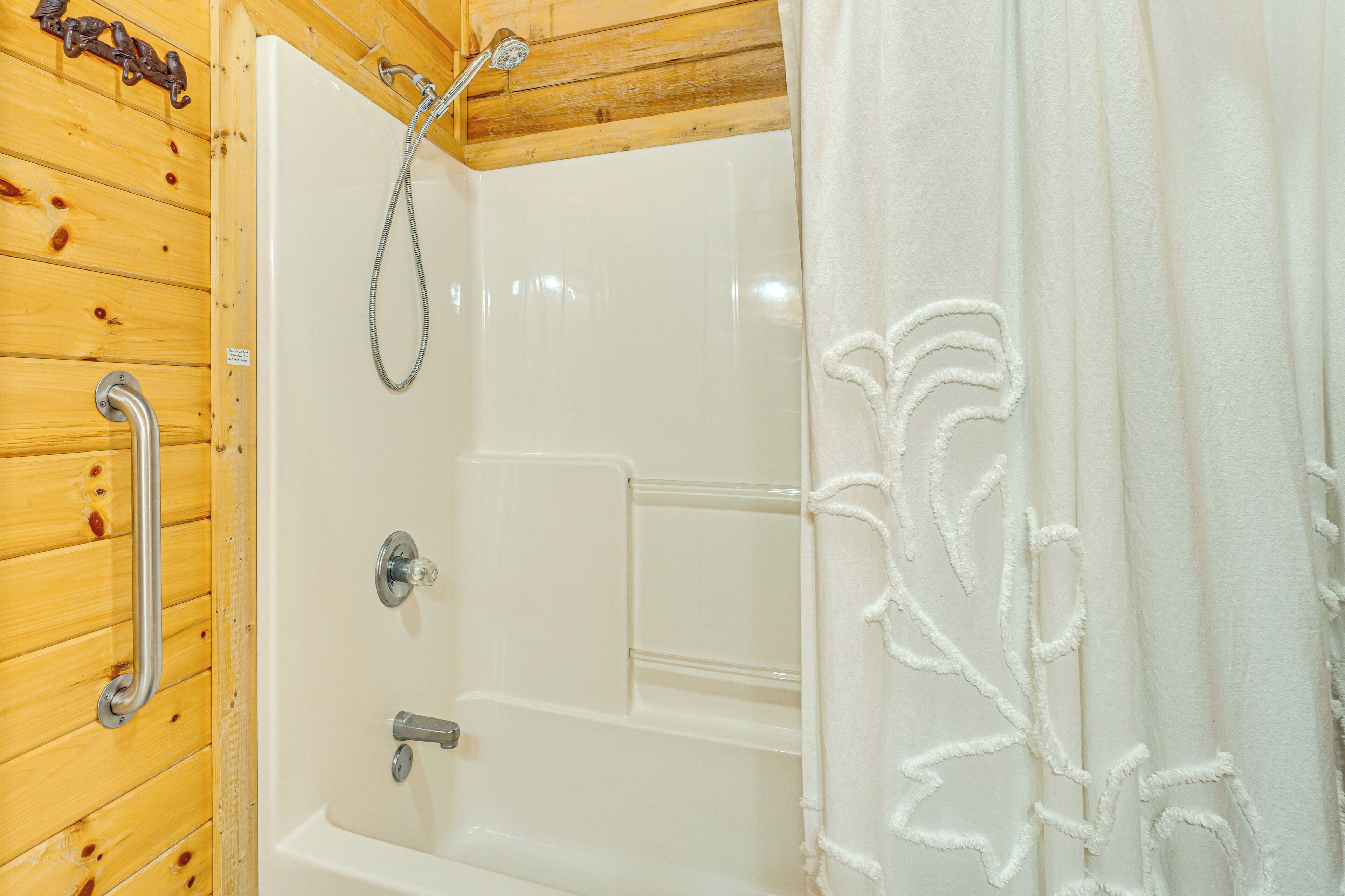 Interno in legno con una vasca da bagno e una doccia bianche, una barra di sostegno in metallo e un tendaggio bianco con motivo floreale.