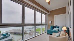 Ein geräumiger Balkon mit großen Fenstern mit Blick auf eine Marina, eine gemütliche Sitzecke mit einem aquamarinfarbenen Sofa und einer Lampe sowie ein Deckenventilator aus Holz.
