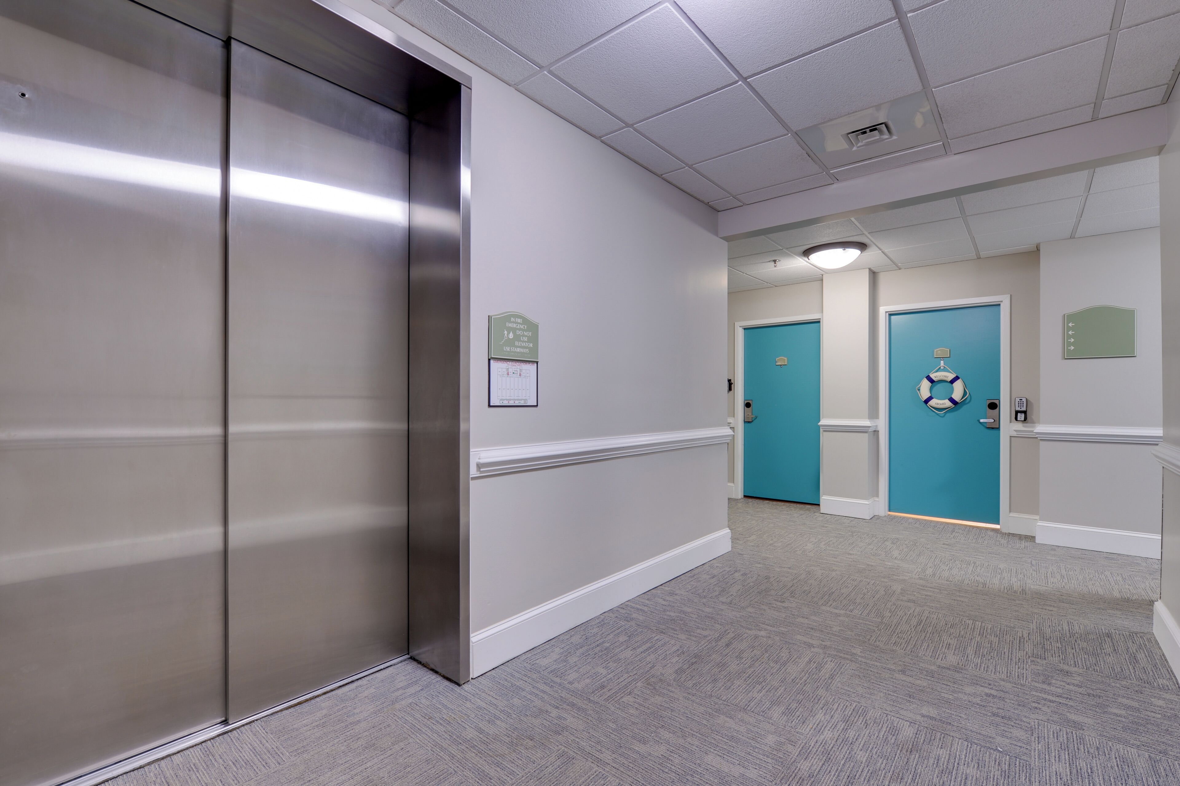 A hallway with a metallic elevator, teal doors, and a carpeted floor.