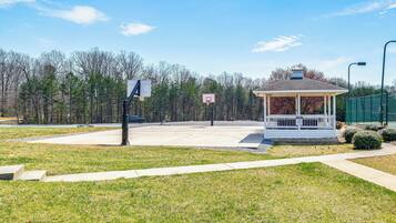 A gazebo with a basketball court in front, surrounded by trees and a clear sky.