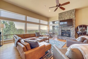 A spacious living room with a stone fireplace, leather sofas, and large windows offering a view of the outdoors. - Panoramic Views: Luxe Payson Paradise! (Payson)