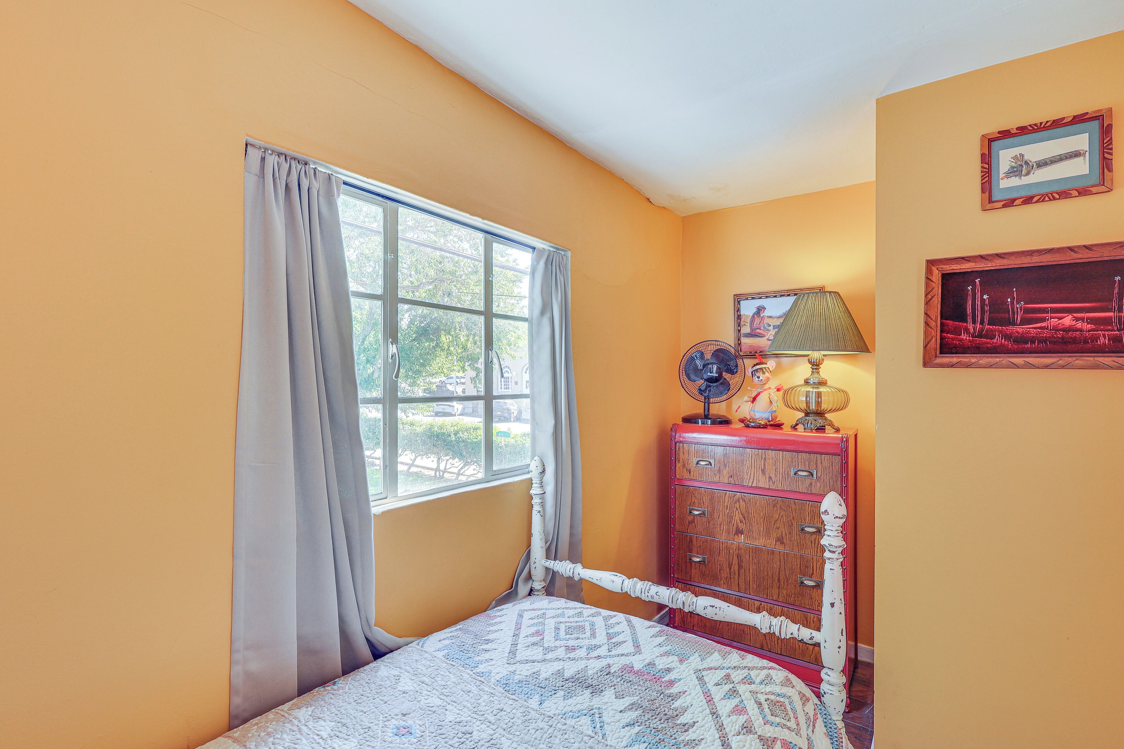 A bedroom with a single bed, a dresser, a window with curtains, and a lamp.