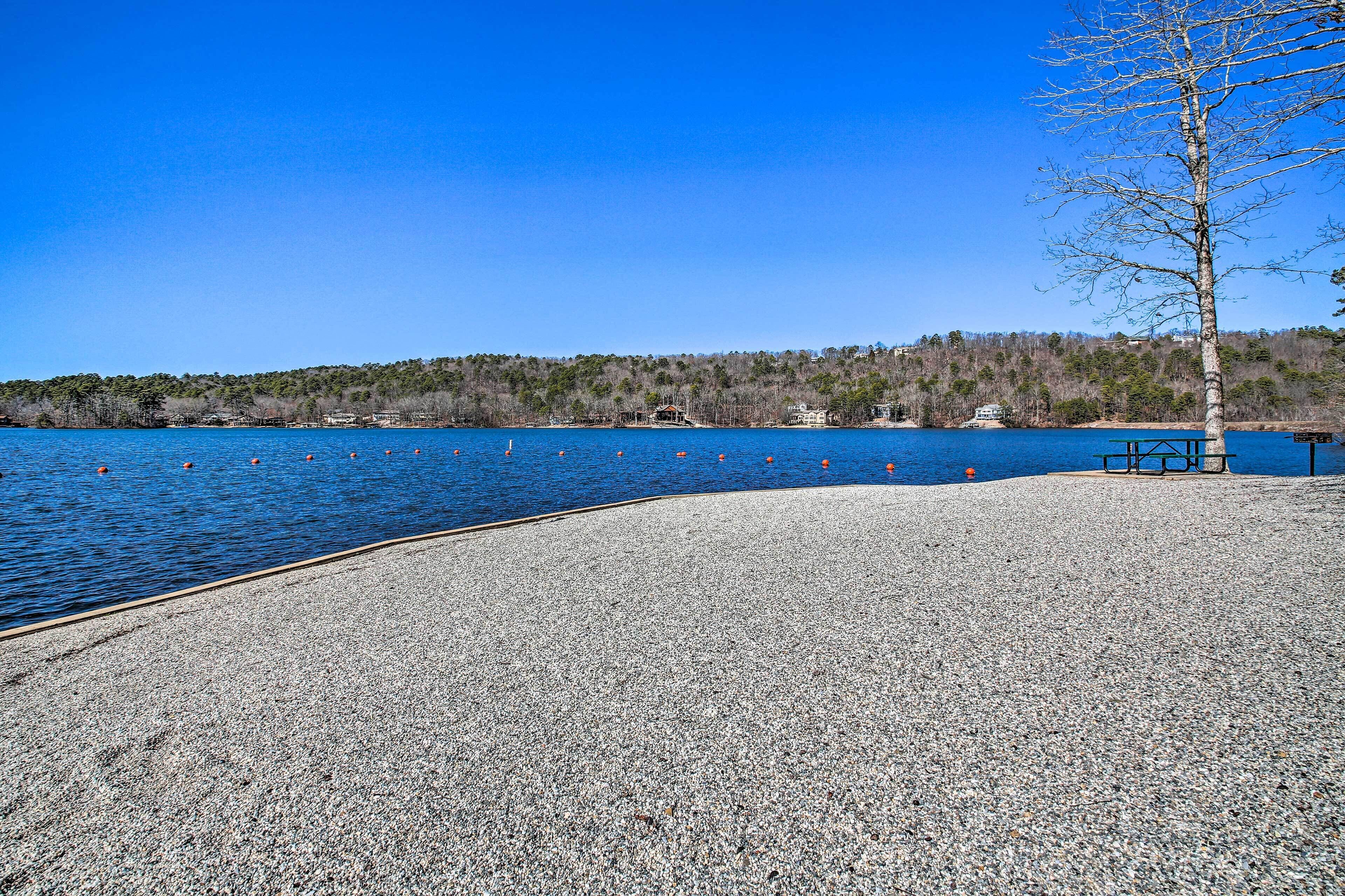 A pebbled beach area with a clear blue sky, a tree, and a distant shoreline with buildings.