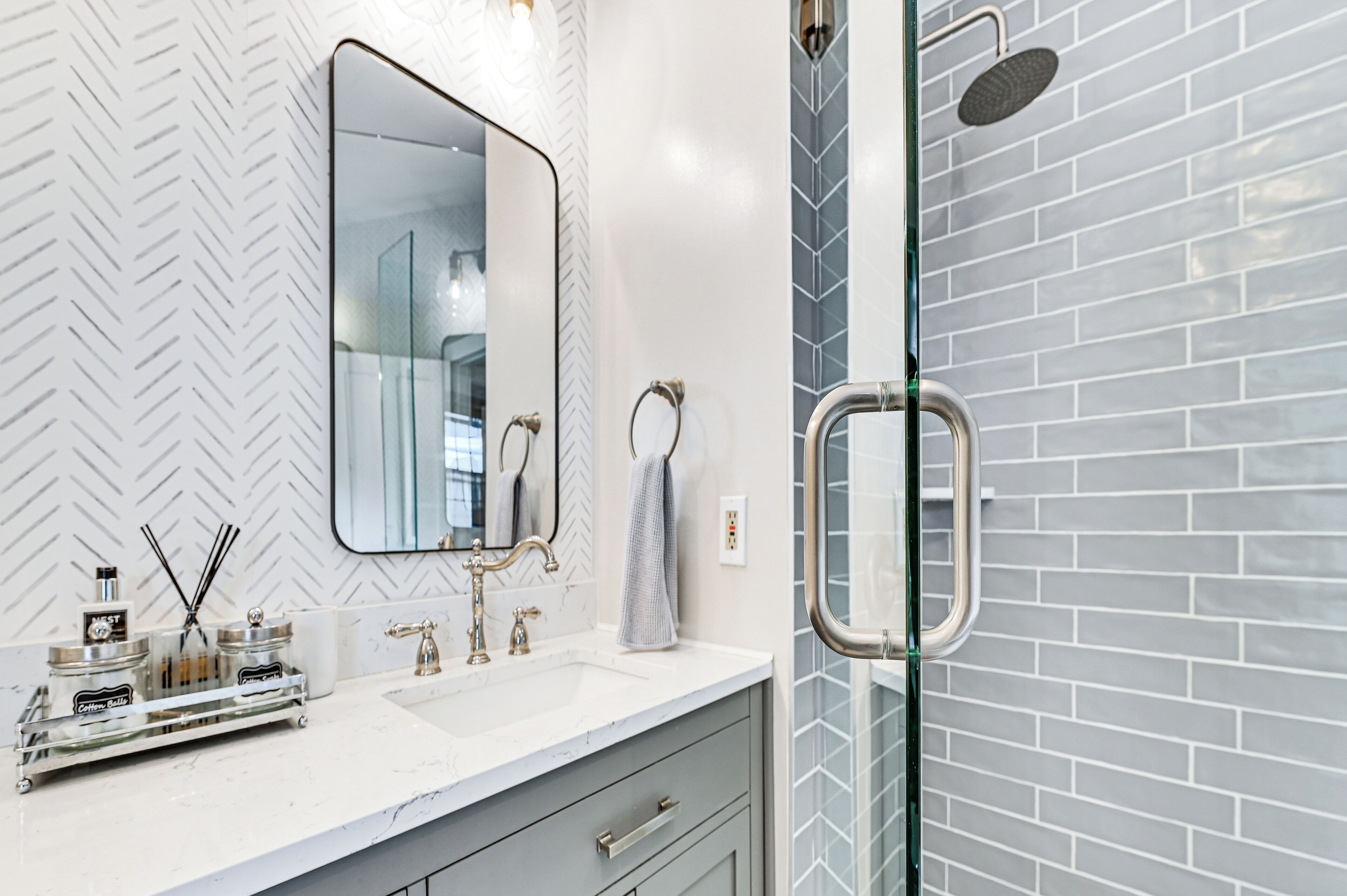Un bagno moderno con una parete bianca a motivi a spina di pesce, un grande specchio, un box doccia in vetro e un lavabo bianco con dispenser di sapone e porta asciugamani.
