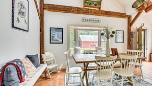 A cozy farmhouse interior with a dining table, white chairs, a bench, and rustic wooden beams.