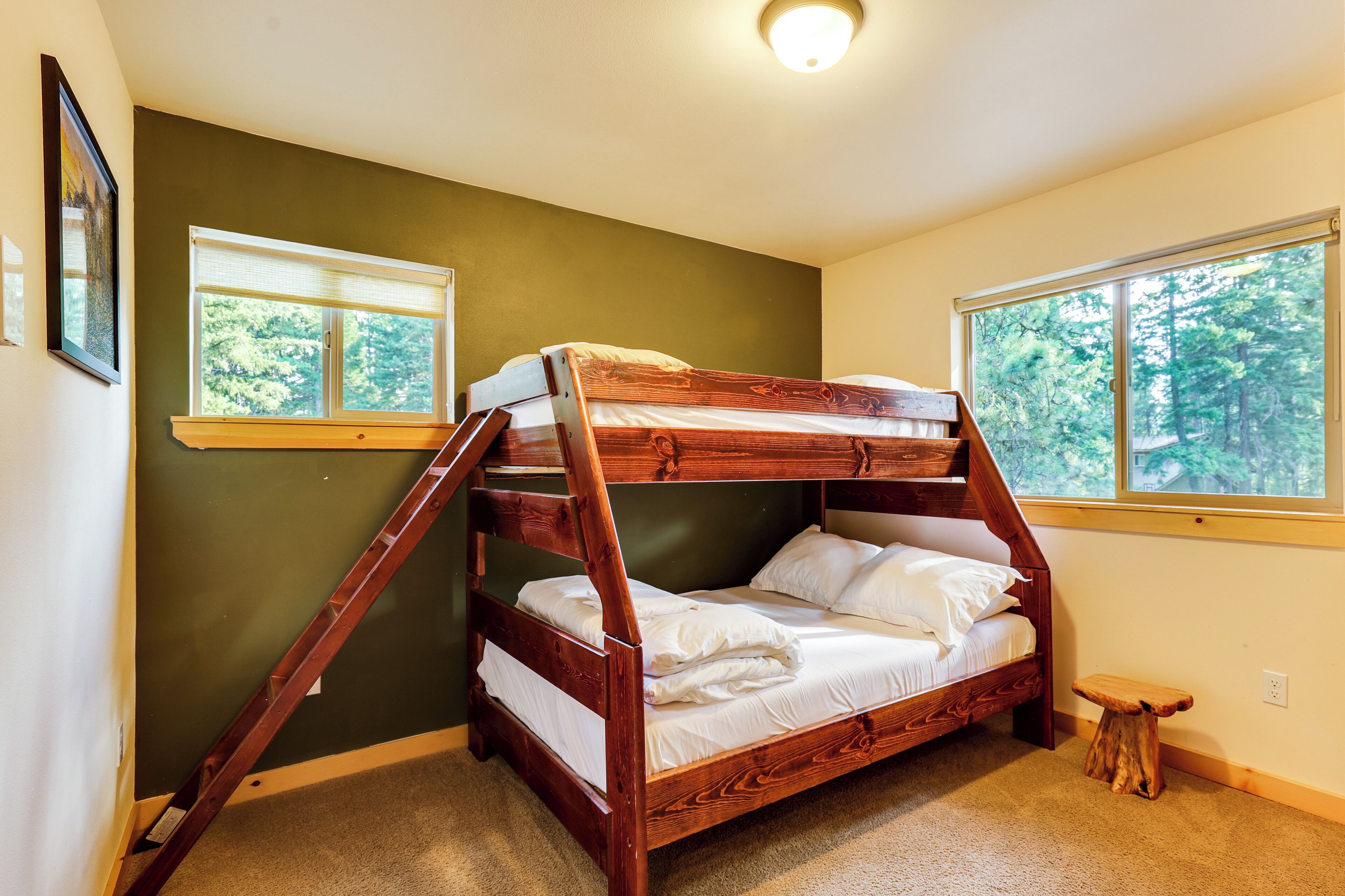 A bunk bed with a ladder, two windows, a small wooden stool, and a framed picture on the wall.