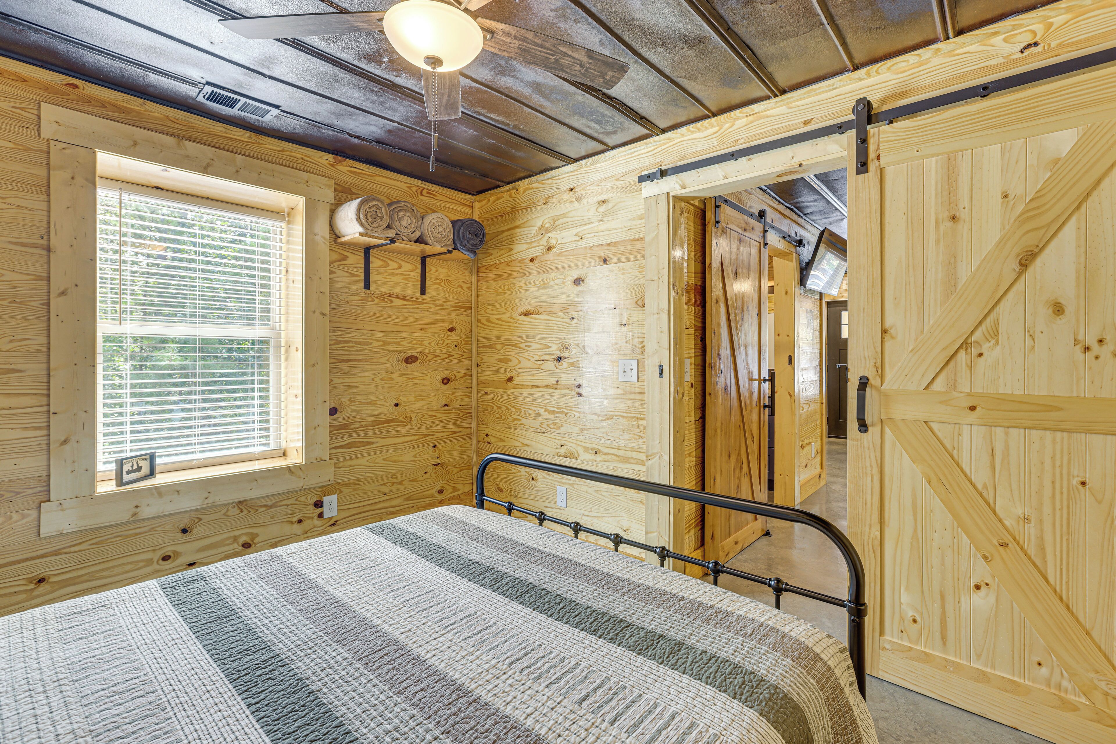 A wooden cabin room with a bed, a window with blinds, a ceiling fan, and a door.