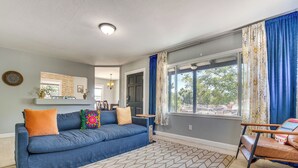Ein Wohnzimmer mit einem blauen Sofa, einem gemusterten Teppich und Blick durch ein Fenster mit Vorhängen.