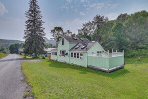 Une maison de couleur vert clair avec une terrasse entourée d’une clôture, une antenne parabolique et un grand arbre à proximité.