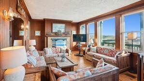 A spacious living room with wooden paneling, a fireplace, and large windows offering a view.