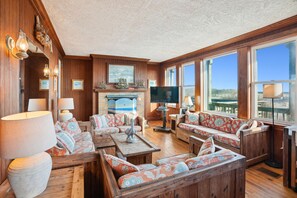 A spacious living room with wooden paneling, a fireplace, and large windows offering a view.