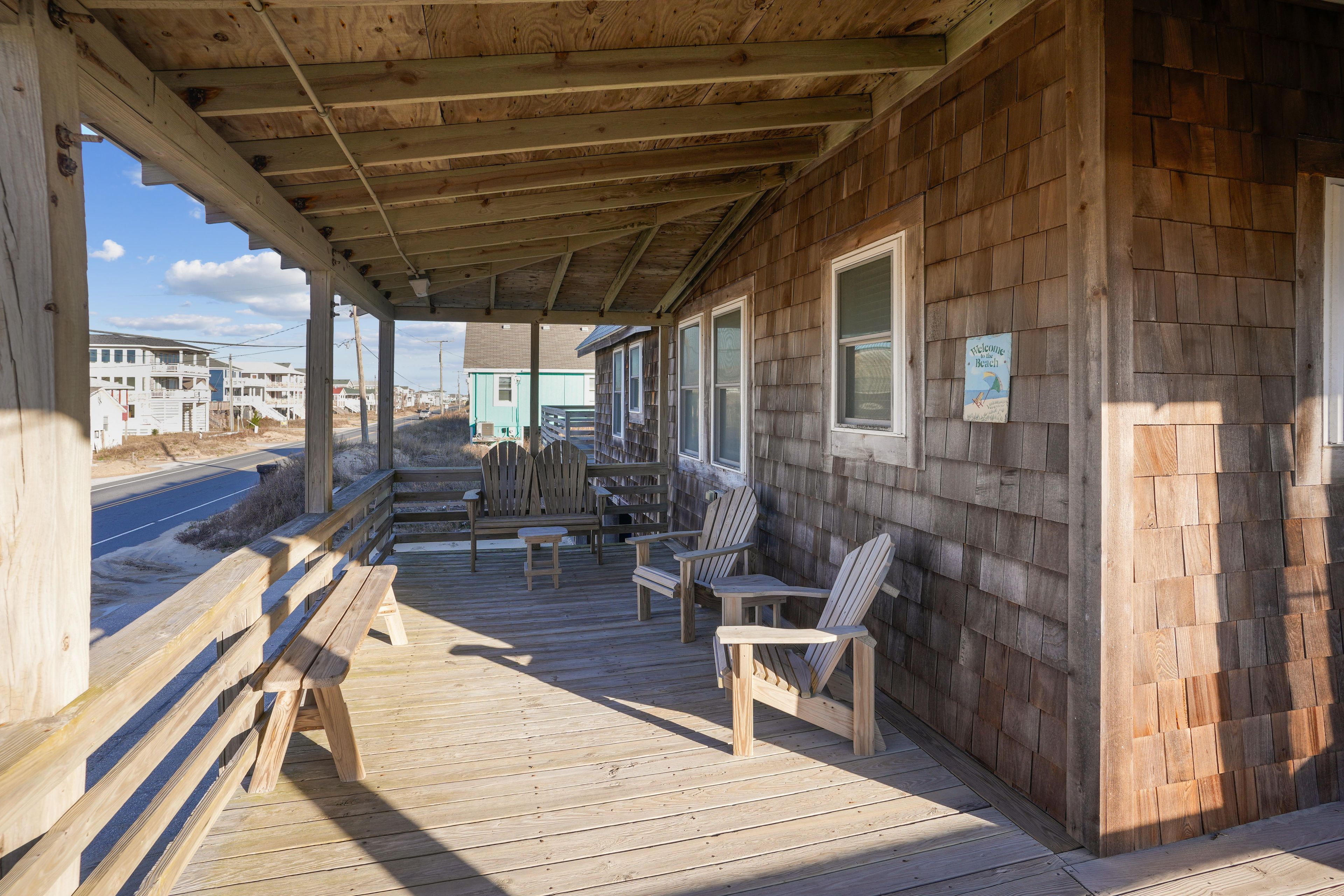 A wooden deck with Adirondack chairs and a bench, overlooking a road with buildings in the background.