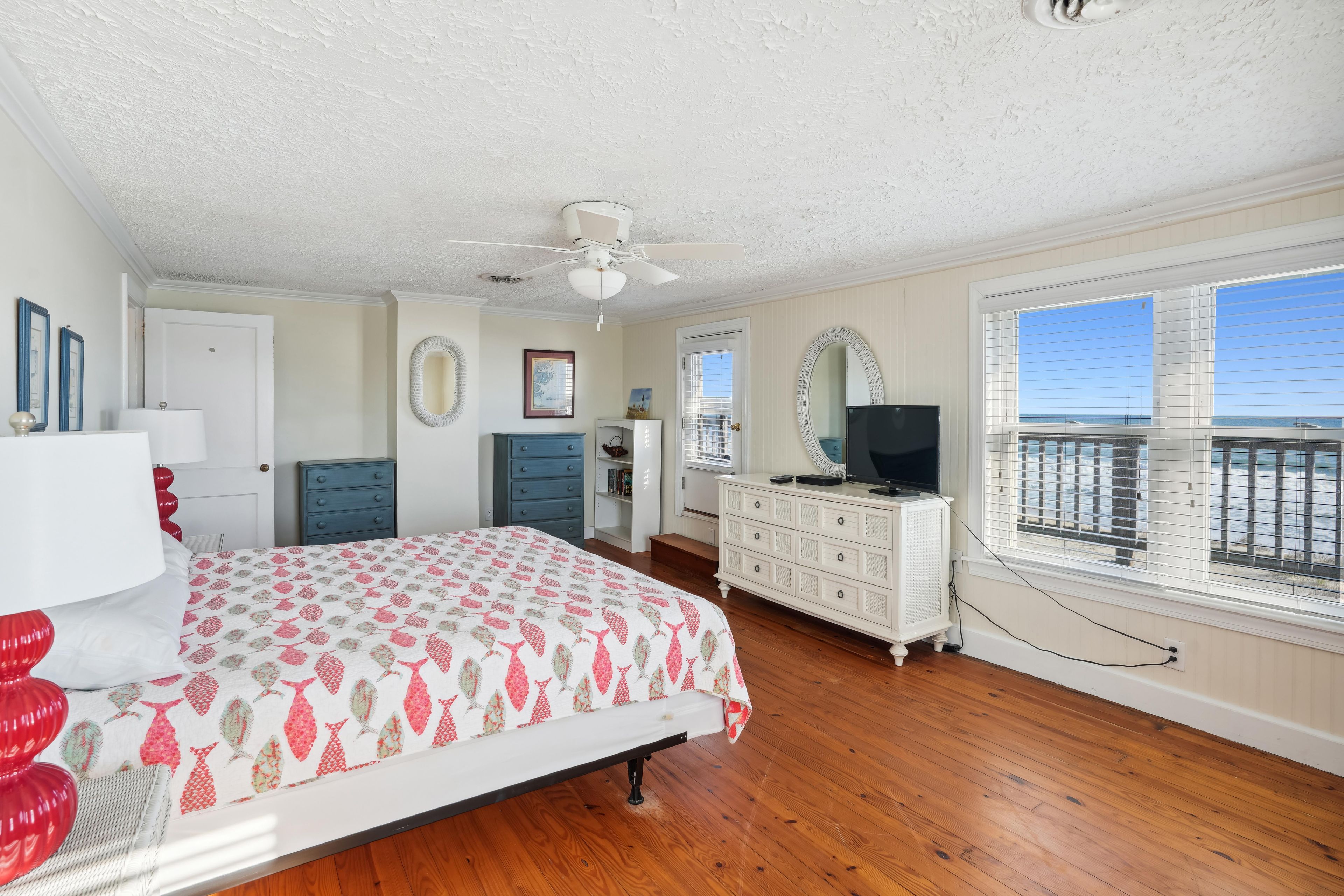 A bedroom with a bed, dresser, and a television. There is a window with a view of the ocean.