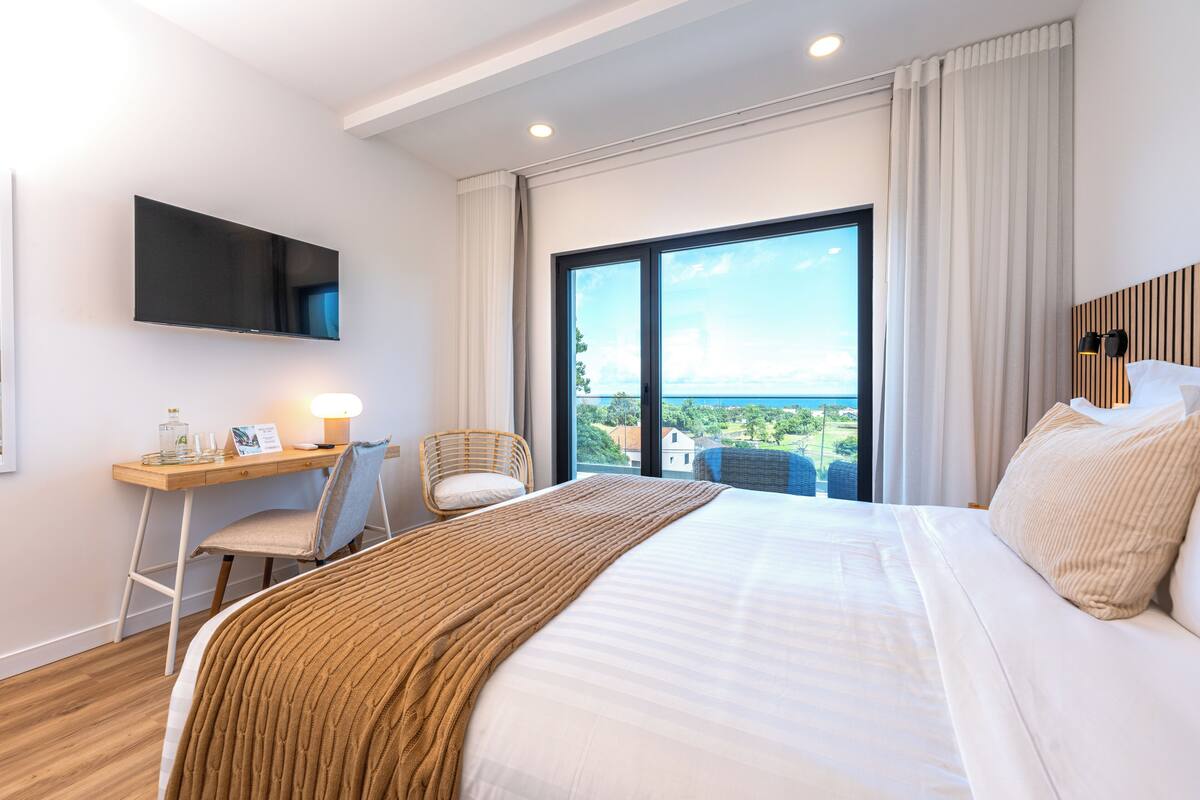 Habitación de hotel con una cama grande, un escritorio con silla, televisor montado en la pared y un ventanal con vistas a la ciudad.