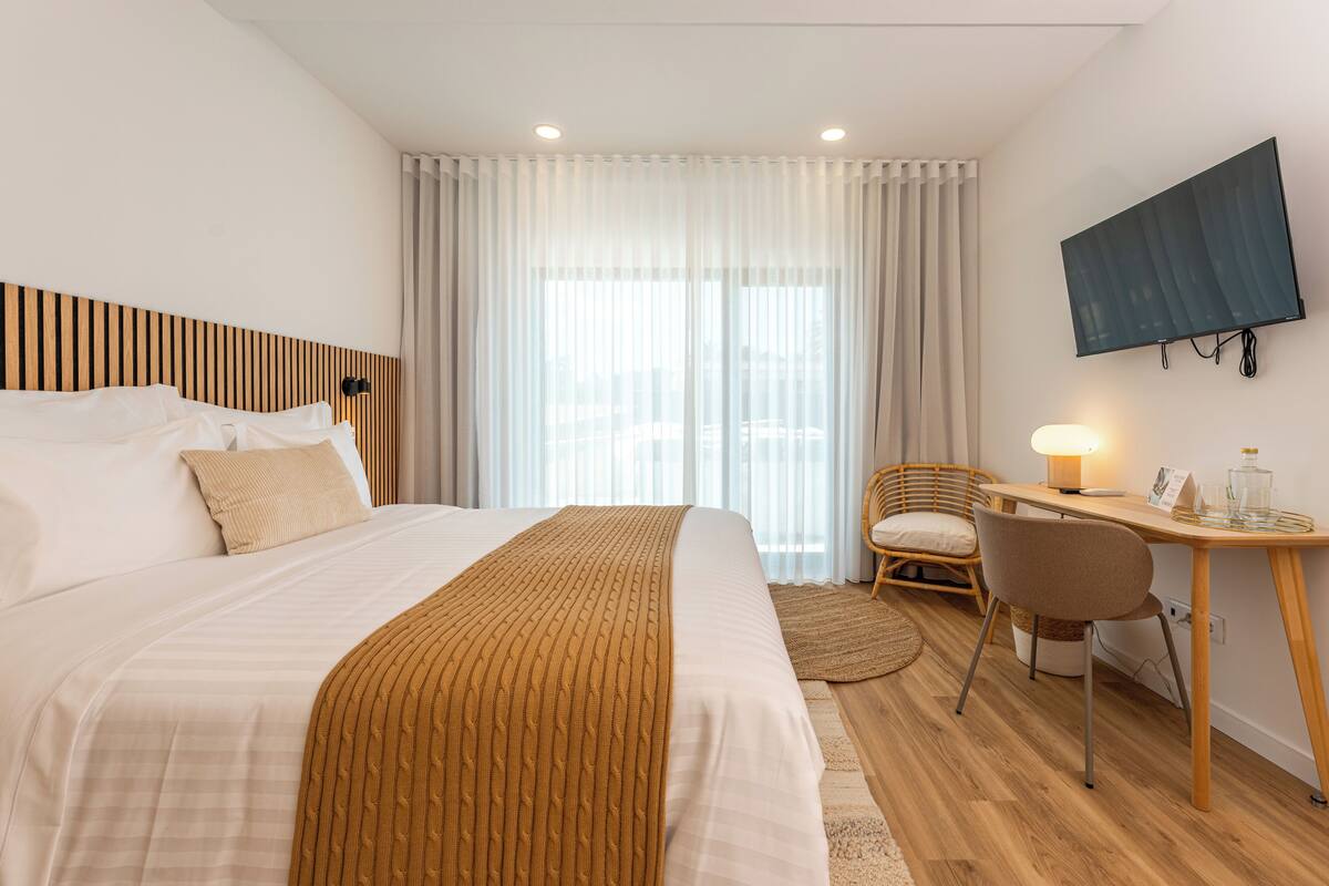 Habitación de hotel con una cama grande, un escritorio con silla, televisión y ventana con cortinas.