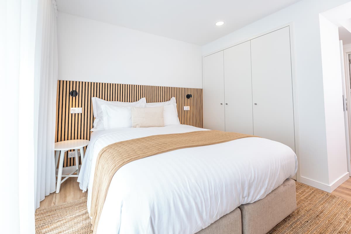 Una habitación de hotel moderna con una cama grande, un cabecero de madera, una mesita pequeña y un armario.
