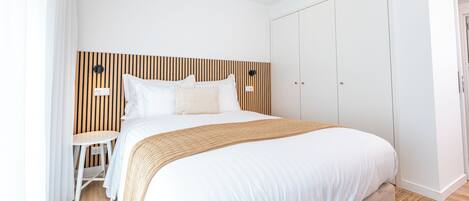 A modern hotel room with a large bed, a wooden headboard, a small table, and a wardrobe.
