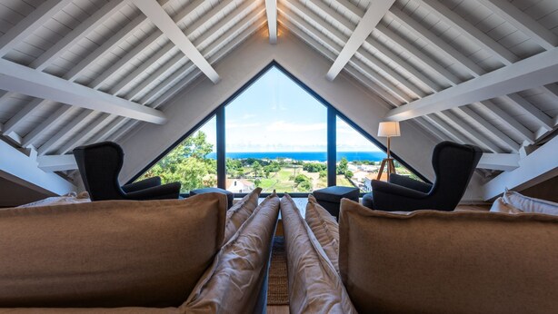 A room with a sloped ceiling, two black chairs, a beige sofa, and a view of the sea. - Ápice - by Azores Green Villas  (Ponta Delgada)