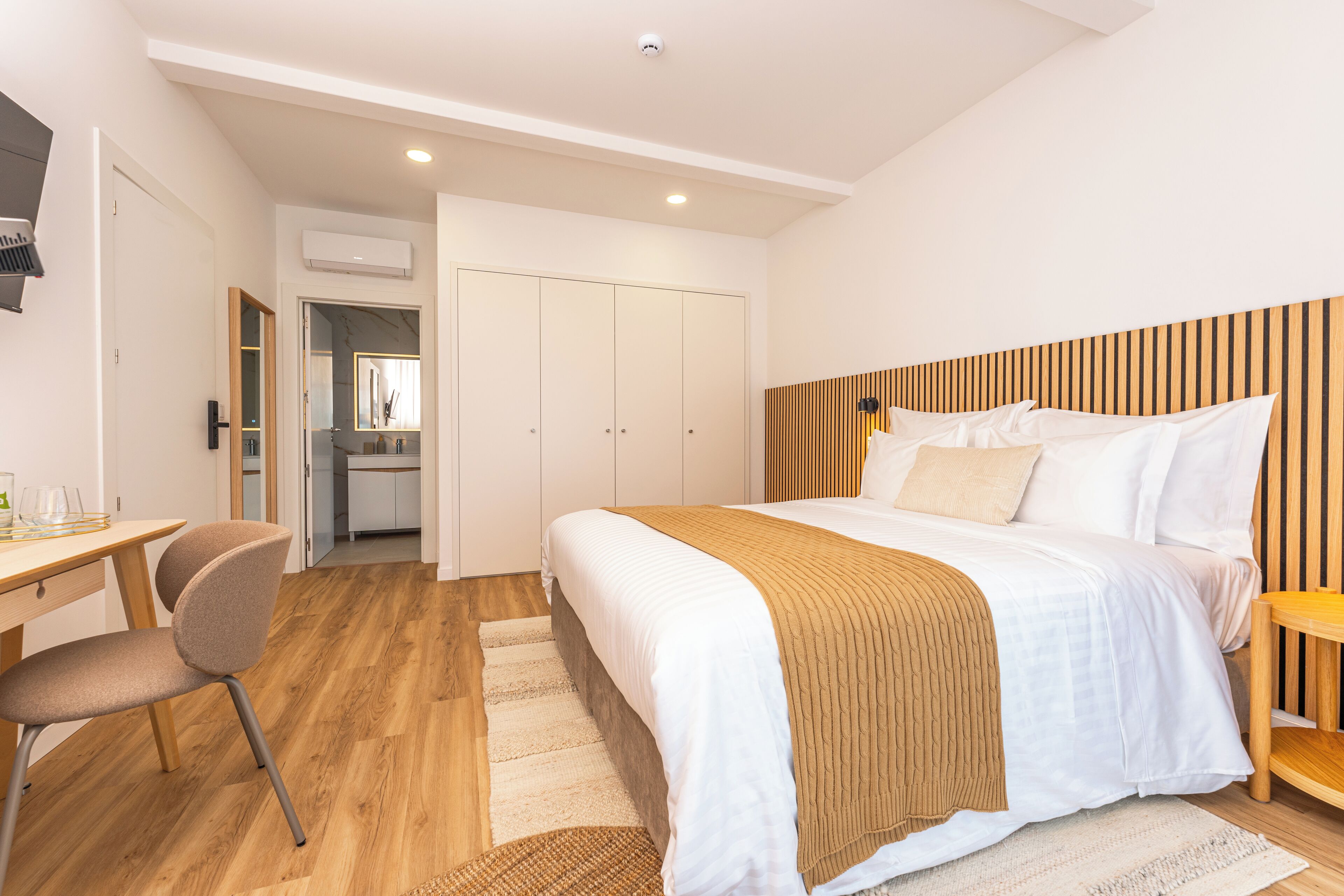 Quarto moderno com uma cama grande, uma escrivaninha e uma cadeira. Há um guarda-roupa e um espelho ao fundo.