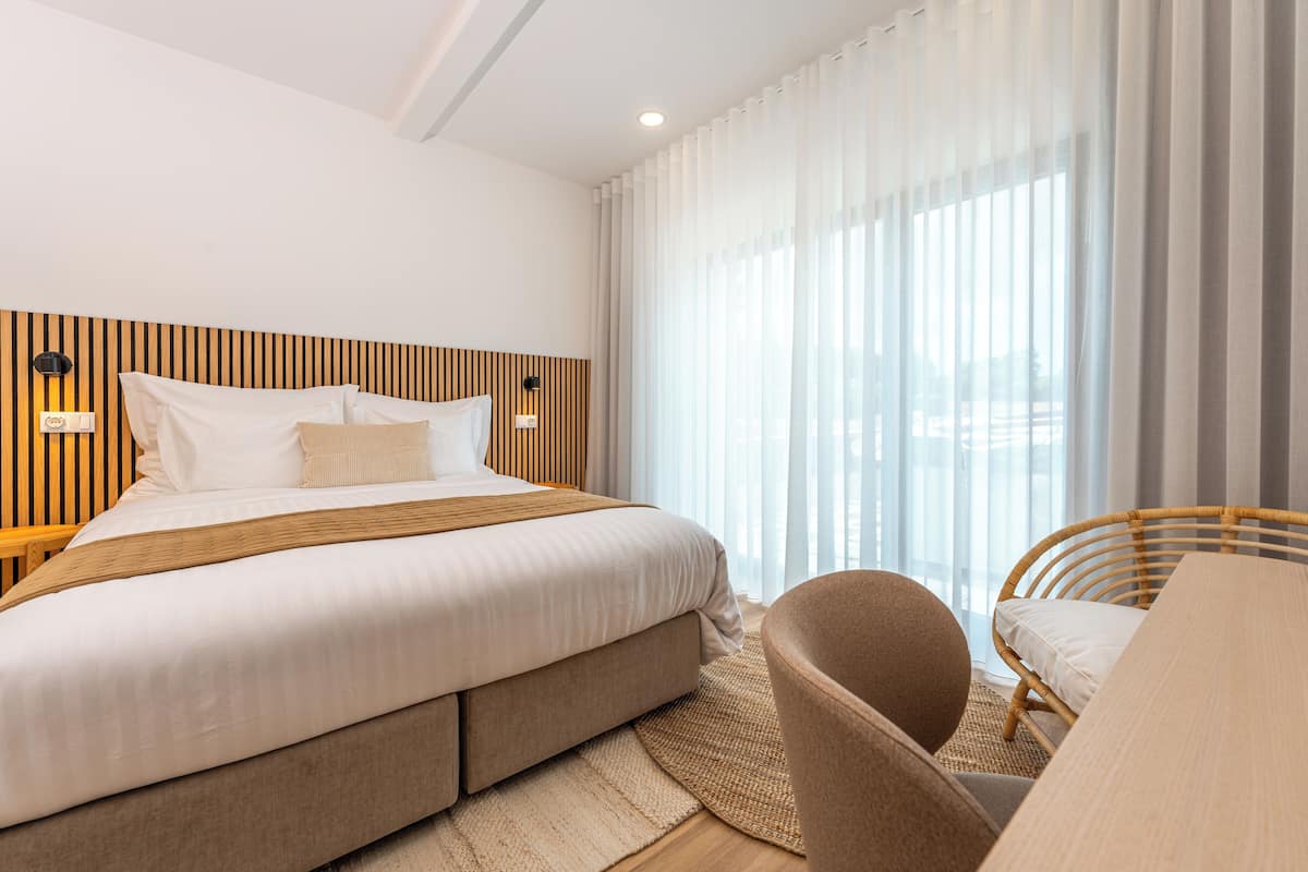 Una habitación de hotel moderna con una cama grande, cabecera de madera, mesita de noche con lámpara, una silla de mimbre y vistas al exterior a través de cortinas translúcidas.