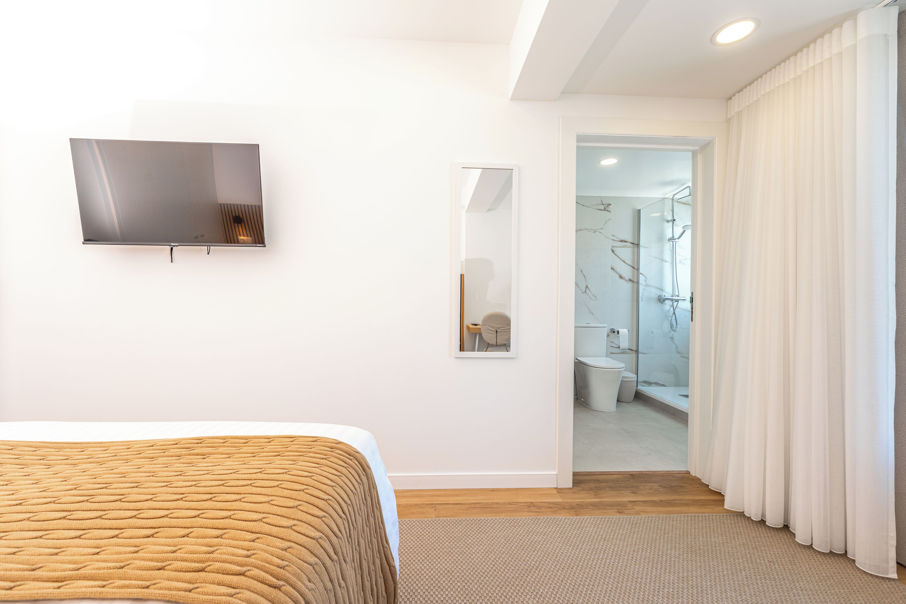 Una habitación de hotel moderna con cama, televisor de pantalla plana, espejo y baño visible a través de una puerta abierta.