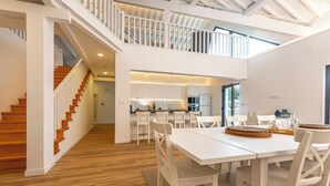A spacious, modern kitchen with white cabinetry and a central island, a dining area with white tables and chairs, and a staircase leading to an upper level. - Ápice - by Azores Green Villas  (Ponta Delgada)