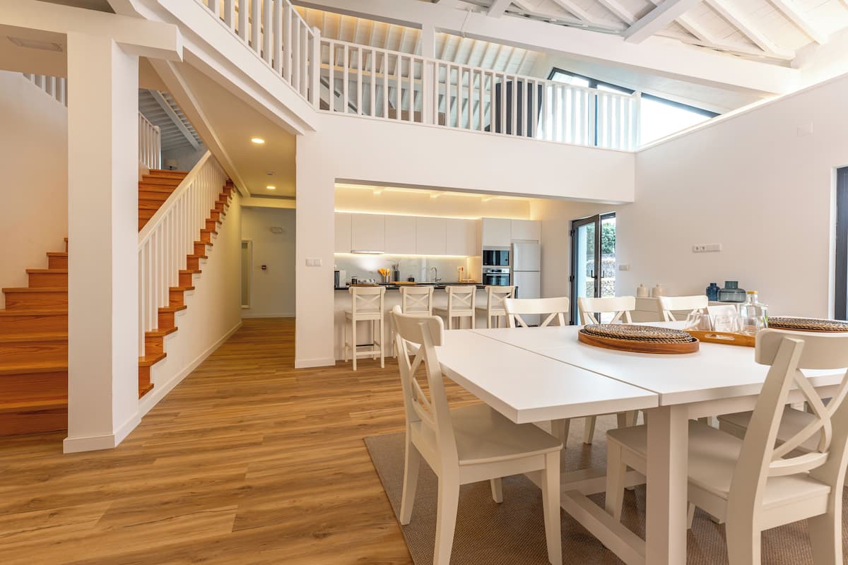 Una cocina amplia y moderna con armarios blancos y una isla central, un comedor con mesas y sillas blancas, y una escalera que lleva a un nivel superior.