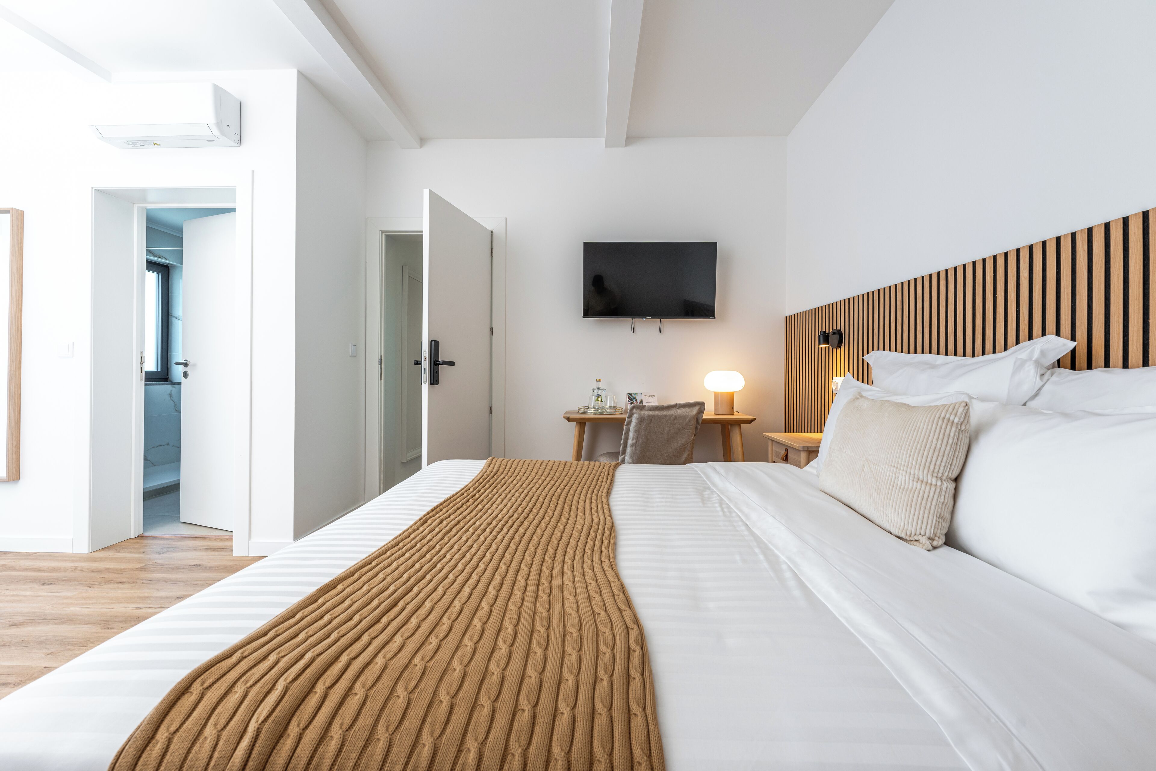 Une chambre d’hôtel moderne avec un grand lit, un téléviseur à écran plat fixé au mur et une tête de lit en bois.