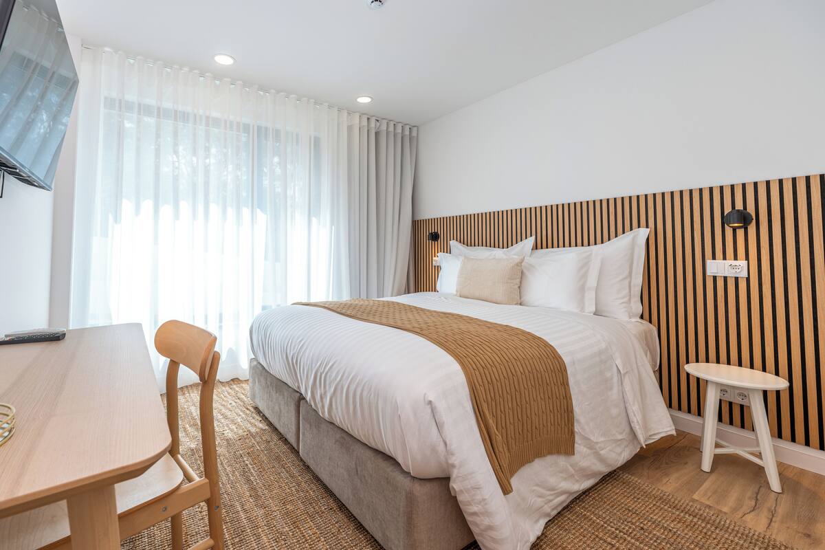Habitación de hotel con cama, escritorio, silla, taburete pequeño y televisor montado en la pared.
