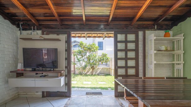A room with a wooden ceiling, a television on a white cabinet, a wooden table, and a view of a garden through an open door. - 3BR House 230m From Beach w/ BBQ & Grg Rgv115 (Bombinhas)