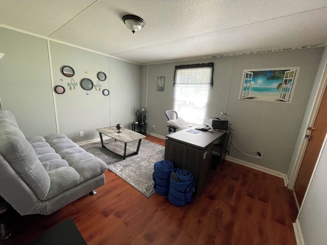 Un espace de vie compact comprenant un canapé gris, une petite table, un bureau avec une chaise et une fenêtre avec des stores.
