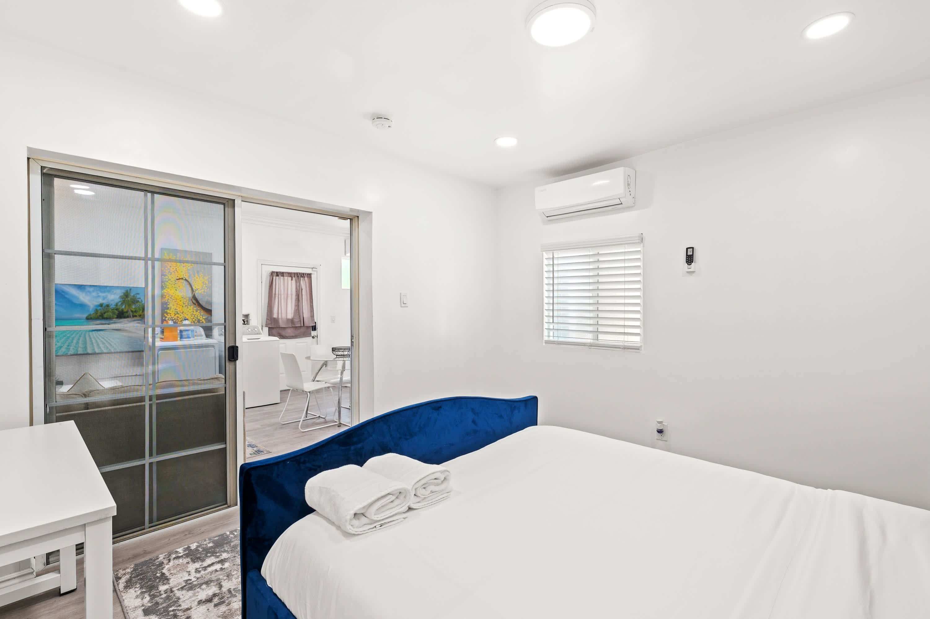 A hotel room with a large sliding glass door leading to a balcony, a bed with blue headboard, a white desk, and a small table.