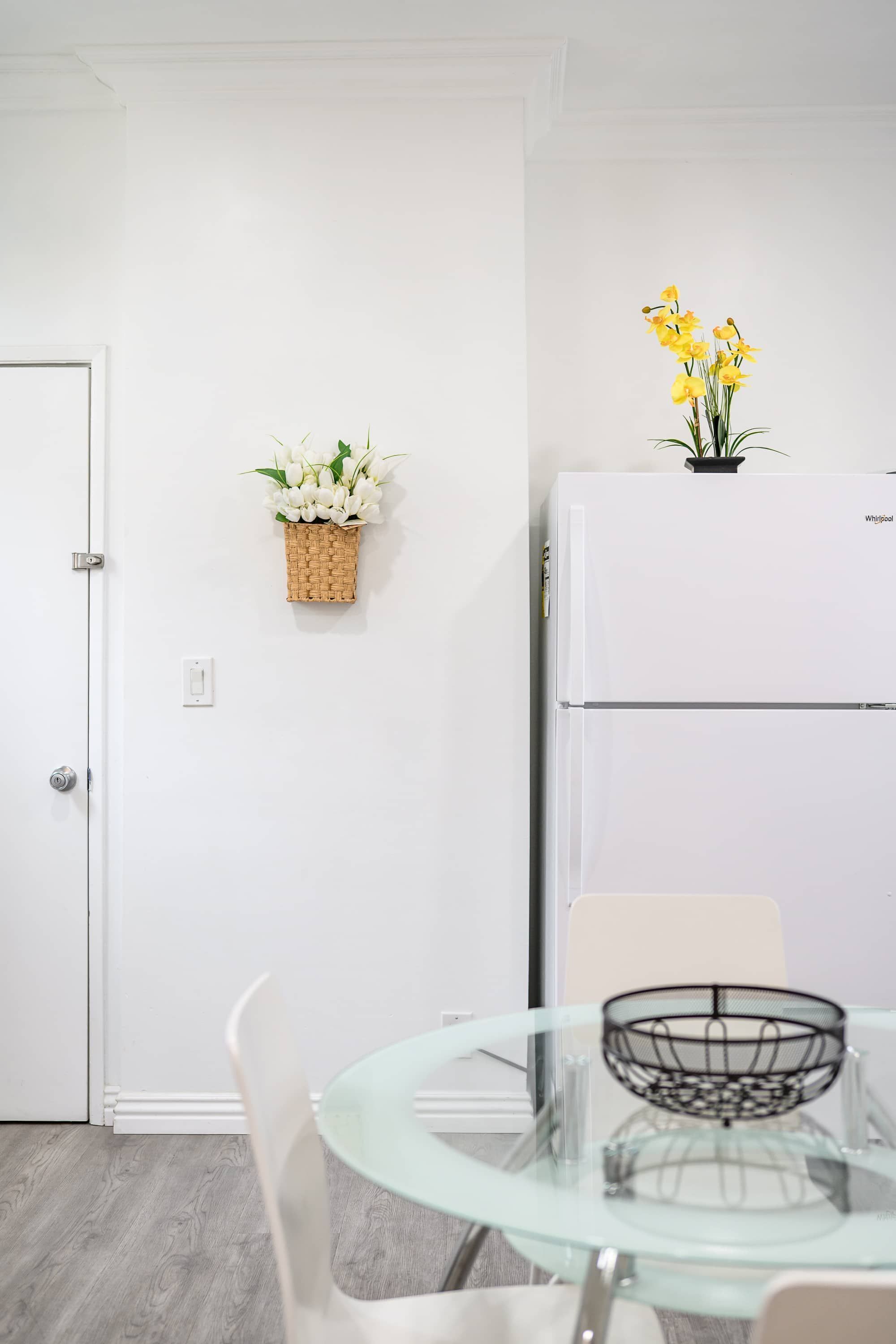 A modern kitchen with a white refrigerator, a glass table with a metal tray, and a basket of flowers on the wall.