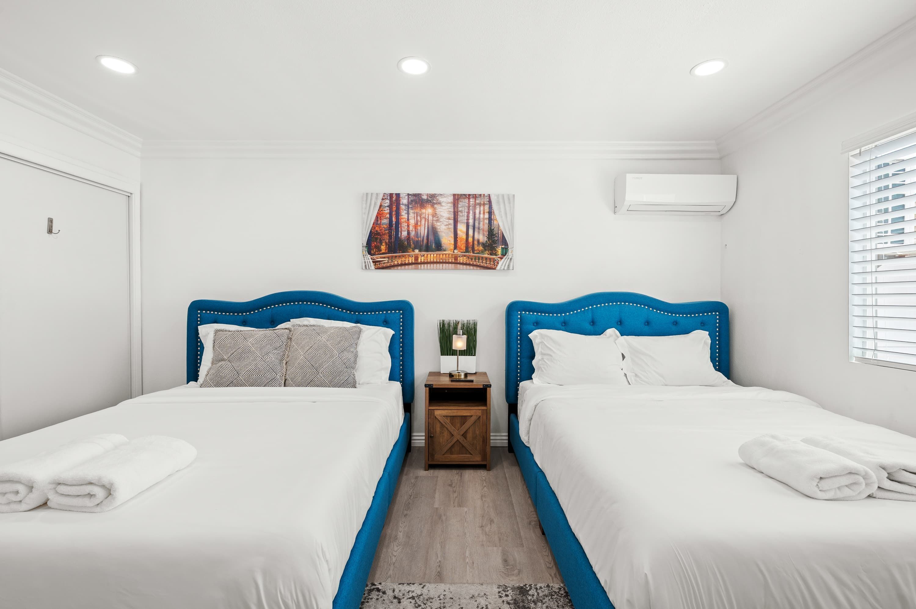 Une chambre d’hôtel avec deux lits, du linge de lit blanc, des têtes de lit bleues, une petite table de chevet en bois, un climatiseur fixé au mur et un tableau représentant un pont.