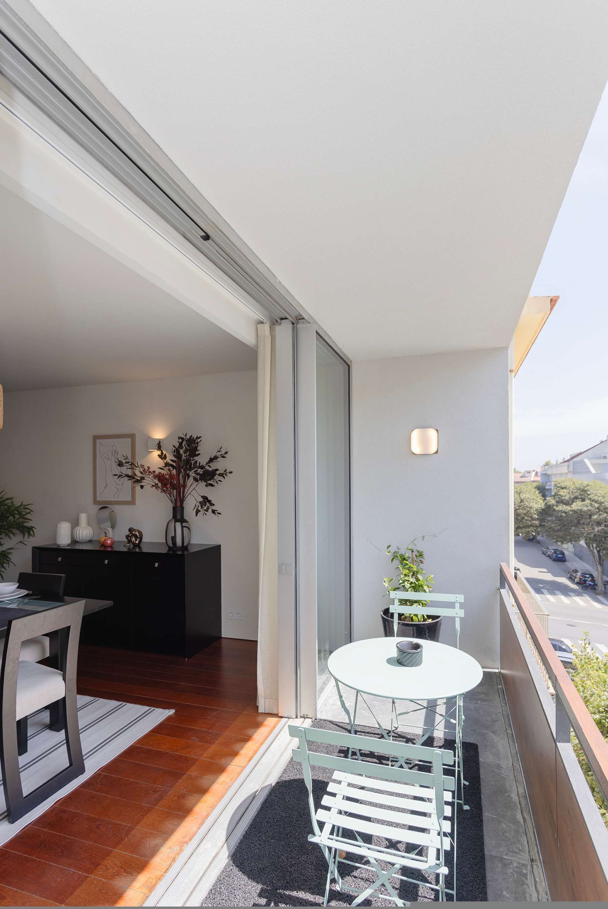A balcony with a dining area, a small table with chairs, a potted plant, and a view of the street.