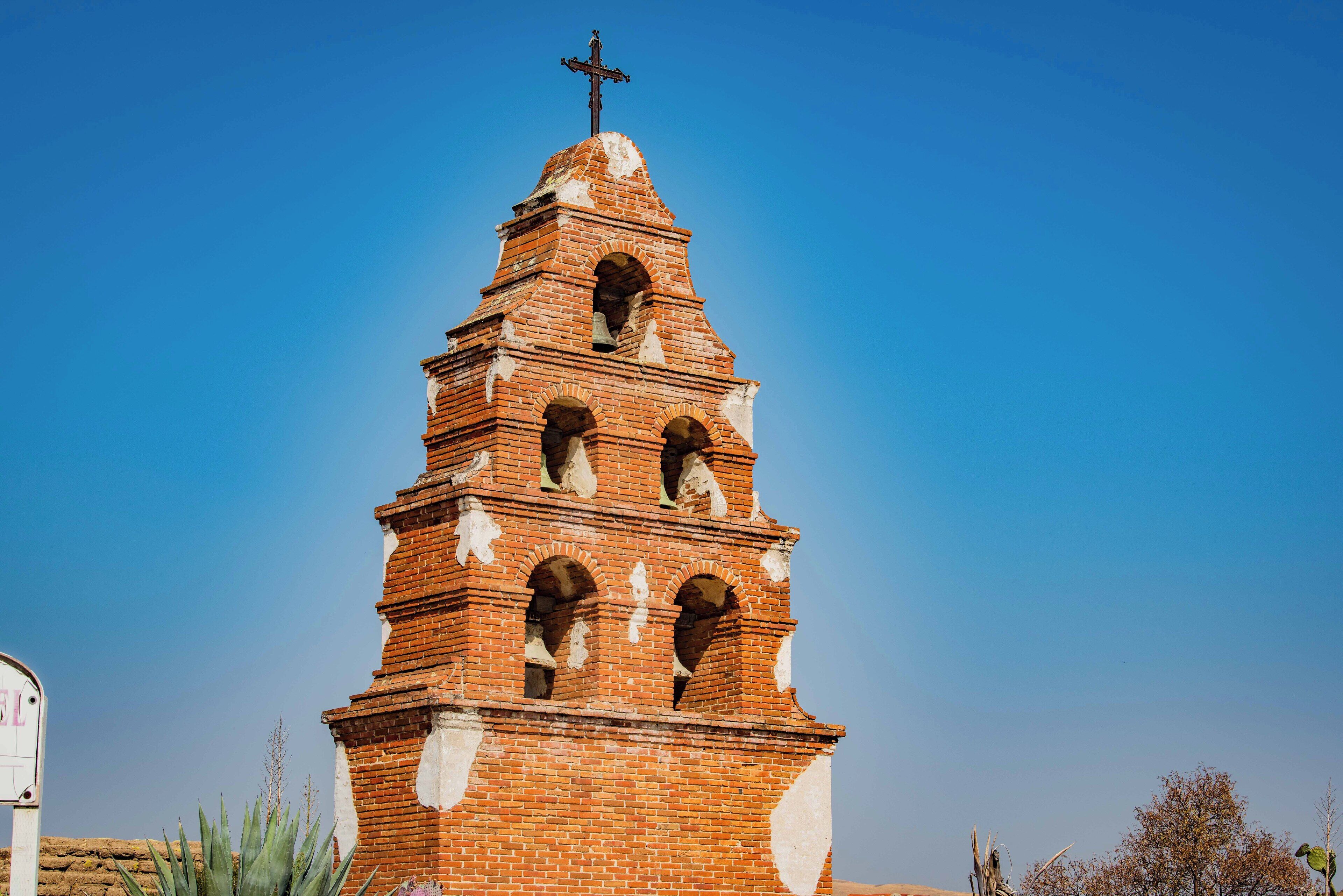 Ein Backsteinturm mit Kreuz auf der Spitze, gewölbten Öffnungen und blauem Himmel im Hintergrund.