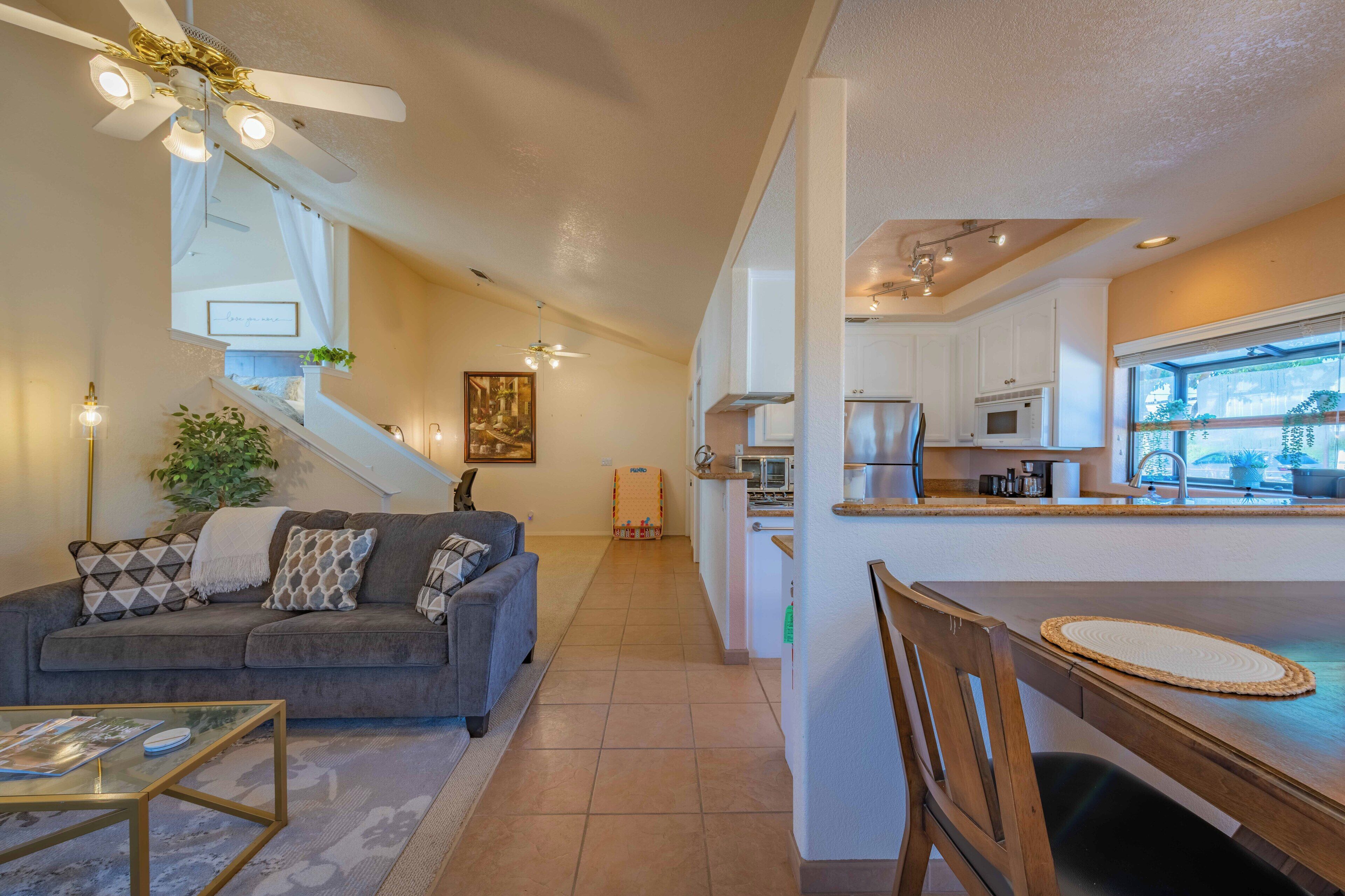 A modern living room with a gray sofa, a glass coffee table, and a dining area with a wooden table and chairs. A kitchen with white cabinets and appliances is visible in the background.