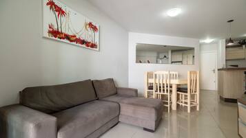 A modern living room with a grey sectional sofa, a dining area with wooden chairs, a kitchen with wooden cabinets, and a large mirror on the wall.