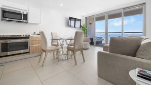 Eine moderne Küche mit Einbauofen und Mikrowelle, ein Essbereich mit Glastisch und Stühlen sowie ein Wohnbereich mit Sofa und Fernseher.