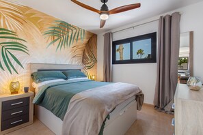 Una camera da letto con un letto, un comodino, una ventola a soffitto, una finestra con tende e un murale raffigurante una foglia tropicale.