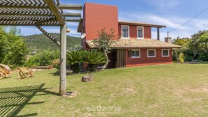 A red house with a tiled roof, a wooden pergola, and a grassy yard with a few plants and rocks. - Comfortable Duplex in Praia do Rosa - SC Rfe000 (Imbituba)
