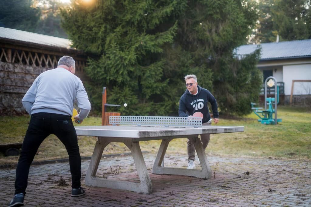Deux personnes jouent au ping-pong sur une surface pavée, à l’extérieur, avec des arbres et un bâtiment en arrière-plan.