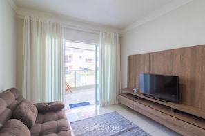 A modern living room with a sofa, a flat-screen TV on a wooden stand, and a sliding glass door leading to an outdoor area. - Beaut apt in Praia do Forte, 700m From sea Rbm207 (Cabo Frio)