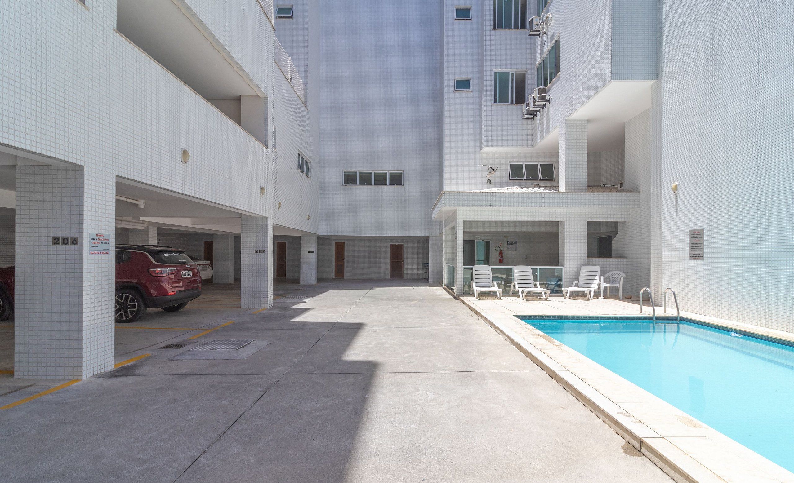 A modern building with a swimming pool, multiple balconies, and a parking area with a red car.