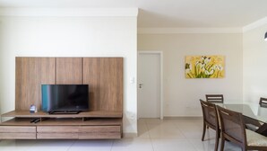 Uma sala moderna com televisão de tela plana em um móvel de madeira, uma mesa de jantar com cadeiras e uma porta que dá para outro cômodo.