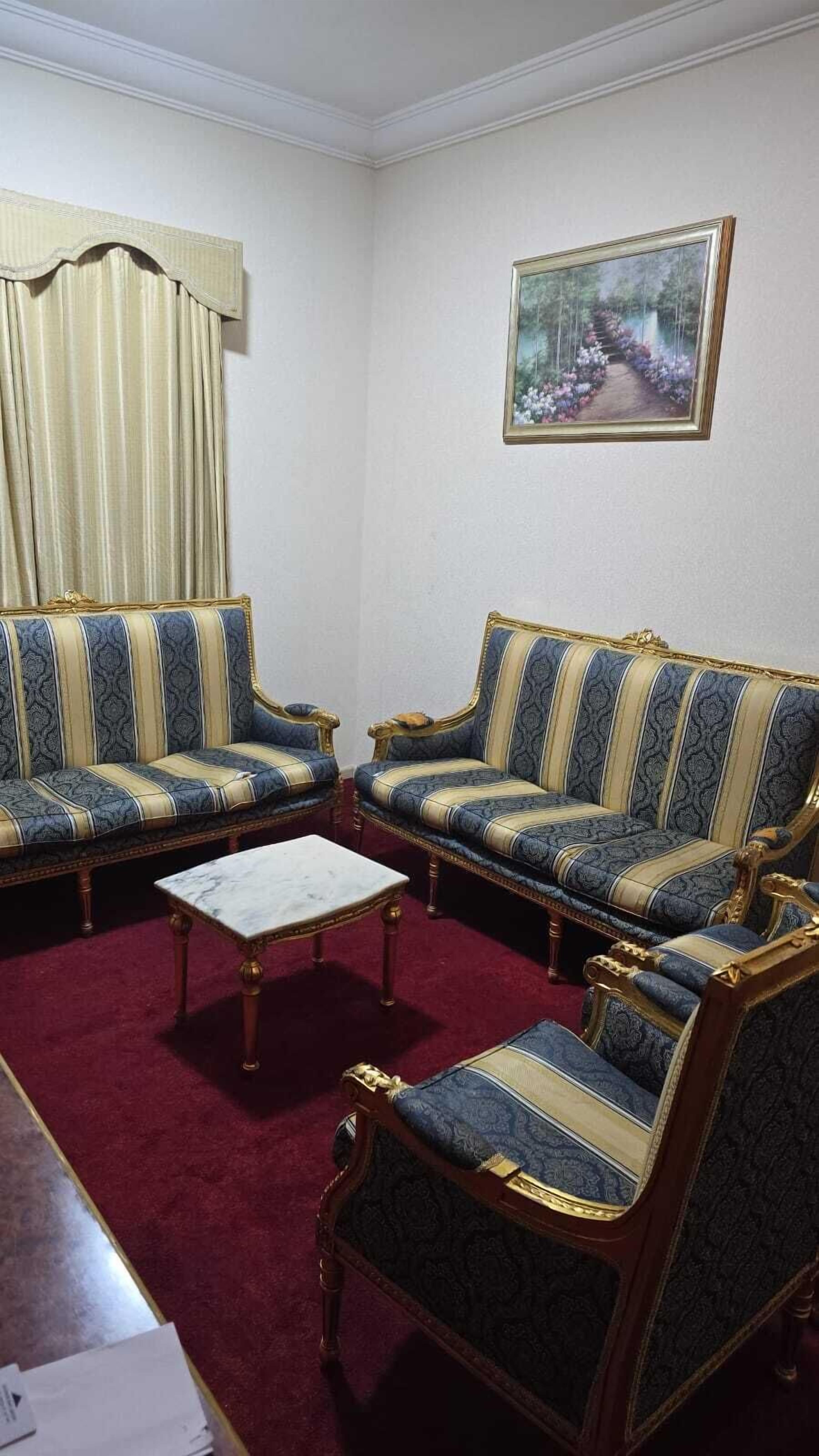 A room with a red carpet, two sofas with gold trim, a white coffee table, and a framed painting on the wall.