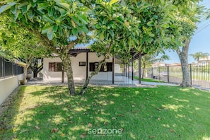 A modern house with a covered patio, a well-maintained lawn, and a tree in the foreground. - 3BR Home w/ BBQ 140m From the Beach Rae0126 (Bombinhas)