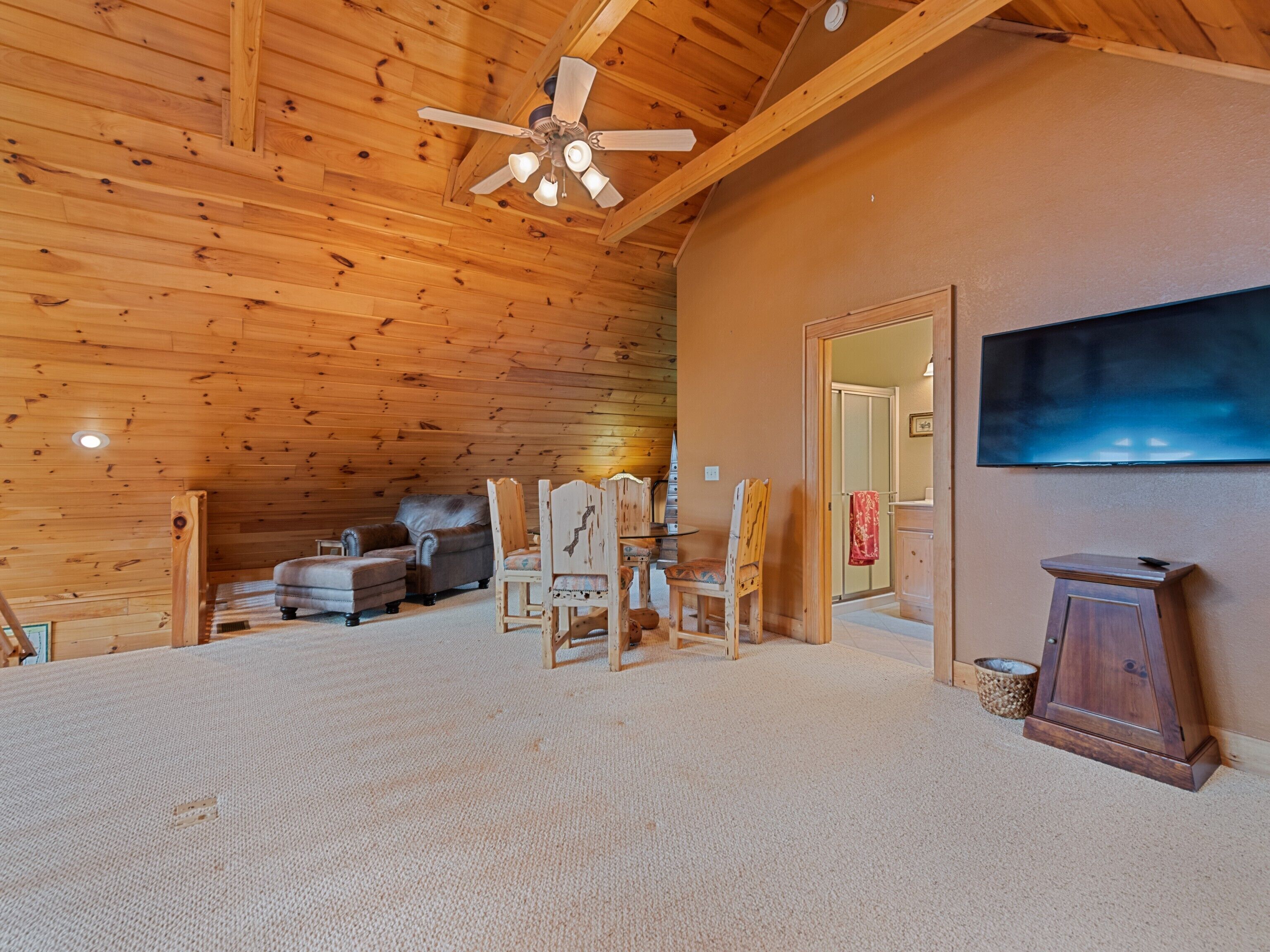Een ruime kamer met een hoge plafond, houten wanden, een plafondventilator, een flatscreen-tv aan de muur, een houten kast en een zithoek met een bank en stoelen.